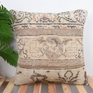Op de afbeelding: Vierkant beige kussen met een vintage tapijtontwerp. Het kussen heeft een centraal patroon met bruine en beige tinten en een gestreepte rand. Het kussen ligt op een houten lattenoppervlak, met een groene plant op de achtergrond.