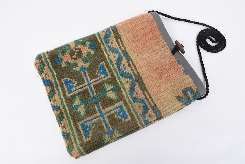 May include: A small, rectangular bag made from a woven rug with a geometric pattern in shades of blue, green, and beige. The bag has a black strap and a wooden toggle closure.