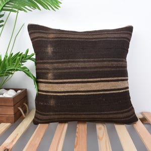 Puede incluir: Un cojín cuadrado con rayas horizontales en tonos marrón y beige. El cojín está hecho de una tela texturizada y se coloca sobre una base de listones de madera. Una planta en maceta es visible en el fondo.