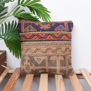 May include: A colorful patterned throw pillow with a geometric design in shades of orange, blue, and brown. The pillow is sitting on a wooden slatted surface.