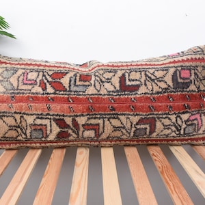 May include: Rectangular beige pillow with a detailed pattern in red, black, and gray. The pillow features a central band of red with a repeating geometric design. The pillow is placed on a wooden slatted surface.