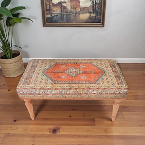 Peut inclure: Un pouf rectangulaire avec un dessus en tapis à motifs orange, bleu et beige. Le pouf a des pieds en bois de couleur claire. Le motif du tapis présente un médaillon central et des détails de bordure complexes. Le pouf mesure environ 120 cm de long.