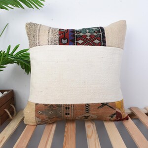 May include: Square decorative pillow with a cream-colored center and patterned top and bottom sections. The top section features a geometric design with red, blue, and green accents. The bottom section has a brown and tan pattern. The pillow is placed on a wooden slatted surface.