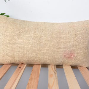 Puede incluir: Una almohada rectangular beige con textura tejida. La almohada está sobre una superficie de madera con listones.