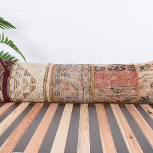 May include: A long, rectangular pillow with a patchwork design. The pillow is made of different fabrics with various patterns and colors, including beige, brown, red, and blue. The pillow is lying on a wooden slatted surface.