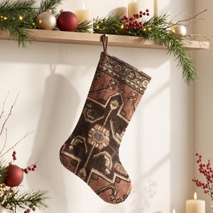 May include: A Christmas stocking featuring a woven rug-inspired pattern in shades of dark brown, muted red, and cream. The stocking has a brown fabric loop at the top for hanging and displays a complex geometric and floral design throughout its surface.