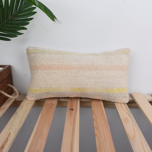 May include: A long, rectangular throw pillow with a cream background and horizontal stripes of yellow and peach. The pillow is sitting on a wooden slatted surface.