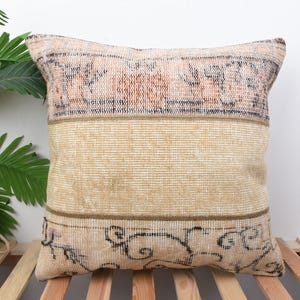 May include: A square decorative pillow with a vintage rug design. The pillow features a beige center with a patterned top and bottom in shades of peach, black, and beige. The pillow is placed on a wooden slatted surface.