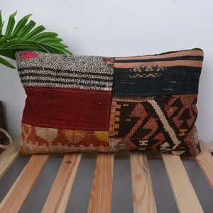 Könnte beinhalten: Rechteckiges Patchwork-Kissen mit einer Vielzahl von Texturen und Farben. Das Kissen hat Abschnitte aus roten, schwarzen, braunen und beigen Stoffen mit geometrischen und gestreiften Mustern. Das Kissen liegt auf einer Holzlattenoberfläche.