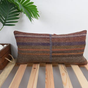 May include: A rectangular pillow with a striped pattern in shades of brown, orange, and purple. The pillow is placed on a wooden slatted surface. A plant in a wooden planter is visible in the background.