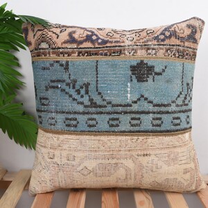 May include: A decorative square pillow with a vintage rug design. The pillow features a light blue central band with black geometric patterns, bordered by beige and brown sections. The pillow is placed on a wooden slatted surface.