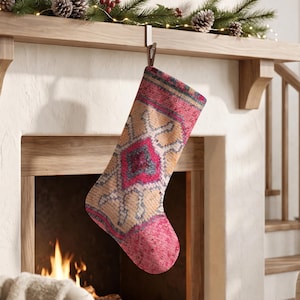 Könnte beinhalten: Ein Weihnachtsstrumpf mit einem lebendigen geometrischen Muster in Pink, Beige und Blau, das an einen gewebten Teppich erinnert. Der Strumpf hat eine strukturierte Stoffoptik mit einem markanten Rautenmotiv in der Mitte und einer einfarbigen Zehenpartie.