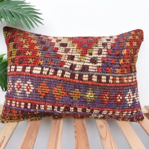 Peut inclure: Coussin rectangulaire décoratif avec un motif tissé. Le coussin présente un mélange de couleurs, notamment du rouge, du violet, de l'orange et du crème. Le coussin est posé sur une surface à lattes de bois.