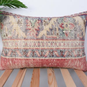 May include: Rectangular decorative pillow with a vintage rug design. The pillow features a beige background with red, blue, and green accents. The pillow is placed on a wooden slatted surface. The pillow's design is reminiscent of traditional Turkish or Persian rugs.