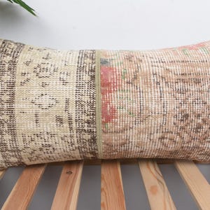 May include: Rectangular decorative pillow with a patchwork design. The pillow features a beige and brown color scheme with floral and geometric patterns. The pillow is placed on a wooden surface.