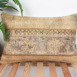 Può includere: Cuscino rettangolare beige con una trama tessuta e una fascia decorativa con motivi geometrici marroni, verdi e viola. Il cuscino è posto su una superficie a doghe di legno.