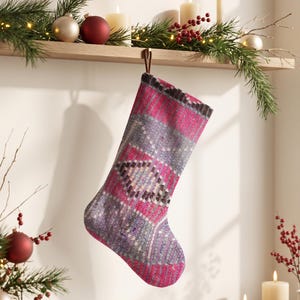 Op de afbeelding: Een kerstkous met een levendig magenta en grijs geweven patroon met geometrische ruitvormen. De kous heeft een bruine ophanglus aan de bovenkant en hangt aan een houten plank die is versierd met kerstdecoratie tegen een effen muur.