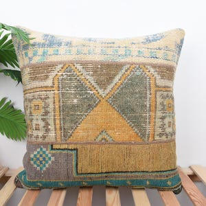 Op de afbeelding: Vierkant decoratief kussen met een geometrisch patroon in beige, bruin, geel en teal. Het kussen heeft een getextureerde, geweven uitstraling en is geplaatst op een houten lattenoppervlak.
