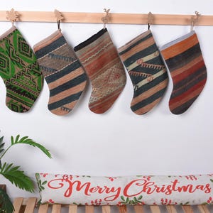 Pode incluir: Cinco meias de Natal coloridas com padrões geométricos penduradas em um suporte de madeira. As meias são feitas de tecido tecido e têm uma variedade de cores, incluindo verde, marrom, vermelho e azul. As meias são decoradas com padrões e desenhos intrincados.