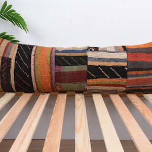 Peut inclure: Un long coussin rectangulaire avec un motif patchwork. Le coussin est fait de tissus de différentes couleurs, notamment le brun, l'orange, le vert et le bleu. Le coussin est posé sur une surface en bois à lattes.