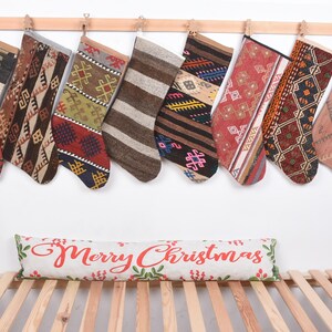 May include: A collection of nine Christmas stockings with various geometric patterns and colors, hanging from a wooden rod. A decorative pillow with "Merry Christmas" text rests below.