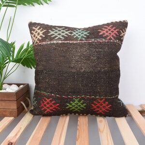 May include: A square, dark brown woven pillow with geometric patterns in red, green, and turquoise. The pillow is placed on a wooden slatted surface, with a potted plant in the background.