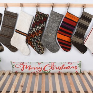 May include: Eight colorful woven Christmas stockings hang on a wooden rail against a white wall. A pillow with the text "Merry Christmas" is on a wooden bed frame below the stockings.