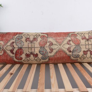May include: A long, rectangular pillow with a red, cream, and blue patterned design. The pillow is made from a woven fabric and has a fringed edge.