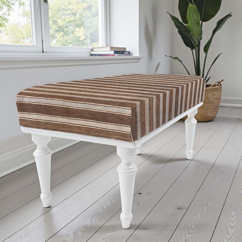 May include: A bench with a brown and beige striped cushion and white legs. The bench is near a window and a large potted plant. The legs are turned and have a classic design. The bench measures approximately 120 cm in length.