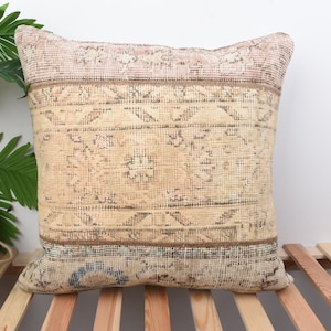 May include: A square decorative pillow with a vintage, distressed look. The pillow features a beige and brown color scheme with intricate geometric patterns. The top and bottom borders have a darker brown hue, while the center has a lighter beige tone. The pillow is placed on a wooden slatted surface.