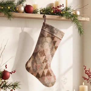 Könnte beinhalten: Ein Weihnachtsstrumpf mit einem verblassten, mehrfarbigen Webmuster in gedeckten Rosa-, Braun-, Grau- und Cremetönen. Der Strumpf verfügt über eine einfache Stoffschlaufe zum Aufhängen und eine texturierte, teppichartige Optik auf der gesamten Oberfläche.