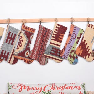Könnte beinhalten: Acht Weihnachtssocken mit verschiedenen geometrischen Mustern in Rot-, Beige-, Grau- und Lilatönen, die an einer Holzstange hängen. Im Vordergrund liegt ein Kissen mit der Aufschrift "Merry Christmas".