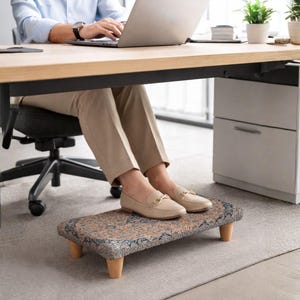 Könnte beinhalten: Ein gemusterter Fußhocker mit Holzbeinen befindet sich unter einem Schreibtisch. Der Fußhocker hat ein beige-blaues Blumenmuster. Eine Person trägt beige Loafer und sitzt an einem Schreibtisch mit einem Laptop.