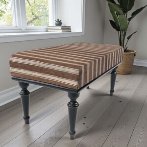 May include: A rectangular bench with a striped brown and beige upholstered top. The bench has a black frame and four dark grey, turned legs. The bench is placed in front of a window and a large potted plant.
