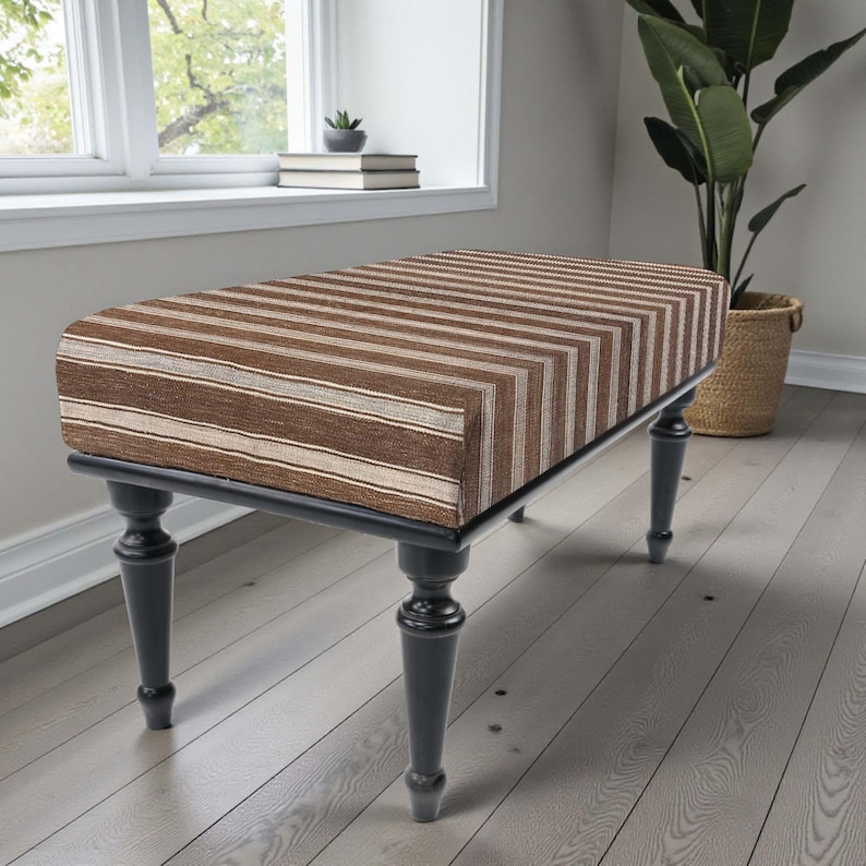 May include: A rectangular bench with a striped brown and beige upholstered top. The bench has a black frame and four dark grey, turned legs. The bench is placed in front of a window and a large potted plant.