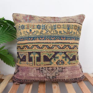 May include: Square decorative pillow with a vintage, patterned design. The pillow features a mix of colors including gold, blue, and purple. The pillow is placed on a wooden slatted surface, with a green plant in the background.