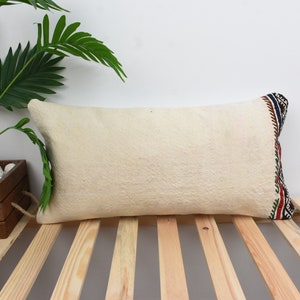 Peut inclure: Un oreiller rectangulaire blanc avec une bordure à motifs colorés. L'oreiller est posé sur une surface en bois à lattes.