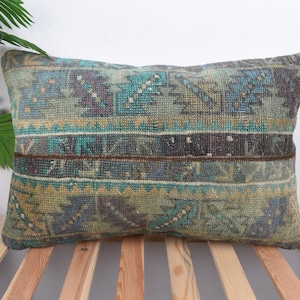 May include: Rectangular cushion with a vintage, distressed appearance. The cushion has a geometric pattern in teal, olive green, and purple, with a brown stripe detail. It is placed on a wooden bed frame.
