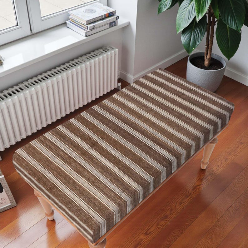 May include: A rectangular bench with a brown and cream striped pattern. The bench has light-coloured wooden legs and a rounded top. The bench is placed on a wooden floor.