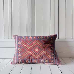 May include: A rectangular pillow with a detailed geometric pattern in shades of red, blue, and beige. The pillow features a central diamond design surrounded by floral motifs and a striped border. It rests on a white wooden floor against a white wall.