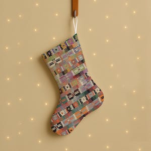 Peut inclure: Une chaussette de Noël avec un motif géométrique coloré, suspendue à un crochet en bois. La chaussette présente un mélange de formes orange, vertes, violettes et noires. Un ruban blanc est utilisé pour suspendre la chaussette.