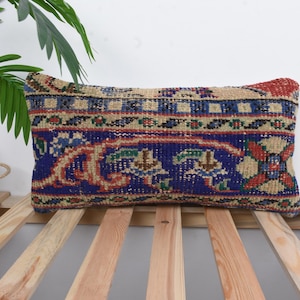 May include: A rectangular cushion with a vintage rug design. The cushion has a blue, red, and beige pattern. It rests on a wooden slatted surface, with a green plant and wooden box in the background.
