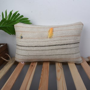 Op de afbeelding: Een wit en beige gestreept kussen met een gele kwast. Het kussen ligt op een houten oppervlak met latten.