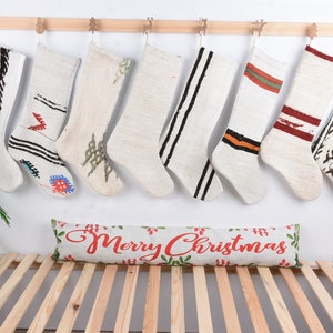 Può includere: Otto calze di Natale bianche con vari motivi e disegni colorati appese a un'asta di legno. Un cuscino bianco con il testo "Buon Natale" in lettere rosse è su una panca di legno.