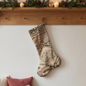 Op de afbeelding: Een kerstkous gemaakt van geweven tapijtstof in vintage-stijl met een gedempt kleurenpalet van oudroze, crème en antracietgrijs. De kous vertoont een ingewikkeld geometrisch patroon met een centraal ruitmotief en een ophanglus.