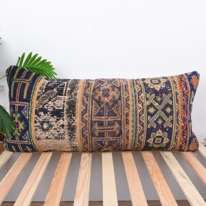 Puede incluir: Cojín decorativo rectangular con diseño patchwork. El cojín presenta una mezcla de patrones en azul marino, beige, naranja y verde. Se coloca sobre una superficie de listones de madera, con vegetación en el fondo.