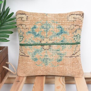 May include: Square beige pillow with a woven design featuring teal and brown patterns. A green band runs horizontally across the center. The pillow is placed on a wooden surface.