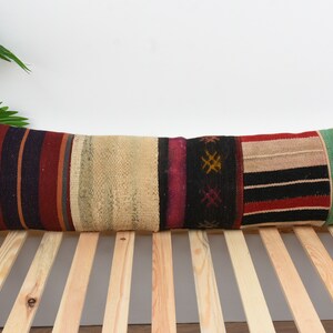 May include: A long, rectangular cushion with a colourful patchwork design. The cushion is made of woven fabric with stripes and geometric patterns in shades of red, black, white, green, and beige.