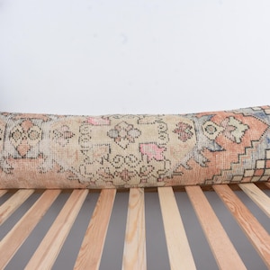 May include: A long, rectangular pillow with a vintage rug design. The pillow features a beige background with intricate patterns in shades of pink, orange, and blue. The pillow is placed on a wooden bed frame, showcasing its decorative appeal.