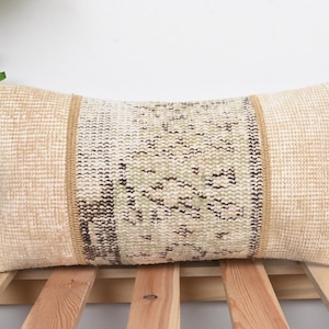 May include: Rectangular beige pillow with a central patterned panel in shades of beige, green, and black. The pillow has a woven texture and is likely made of natural fibers. The design suggests a vintage or bohemian style, suitable for home decor.